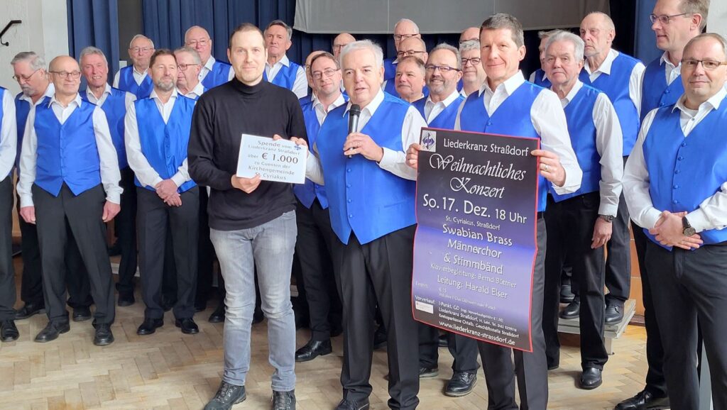 Männerchor in blauen Westen mit 1.000 Euro Scheck und Plakat des Weihnachtskonzertes bei der Scheckübergabe an den Zweiten Vorsitzenden des Kirchengemeinderates
